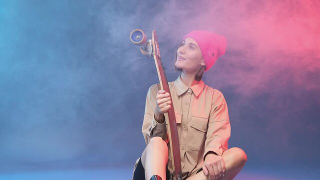 Beautiful young female hipster in stylish clothing and pink hat poses sitting on floor and holding skateboard in colourful and atmospheric background with smoke.