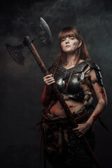 Armed with two handed axe nordic grimy amazon in dark armour with brown hairs poses in dark background with smoke.