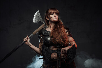 Obraz na płótnie Canvas Barbaric female viking in light armour with brown hairs poses in dark smokey background holding two axes.