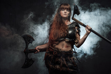 Obraz premium Portrait of beautiful and at the same time dangerous scandinavian woman fighter wielding two axes in dark smokey background.