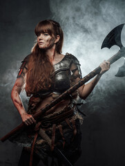 Fototapeta na wymiar Portrait of woman viking wielding two handed axe and dressed in dark light armour in dark foggy background.