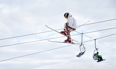 Side view of man skier making jump with cloudy sky and ski lifts on background. Male freerider on skis jumping in the air while sliding down snow-covered slopes. Concept of extreme winter sports