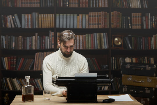 A focused male writer with glasses is typing a novel on a typewriter. A man with a beard in a white sweater using a retro typewriter sitting at a home table in the background of the library