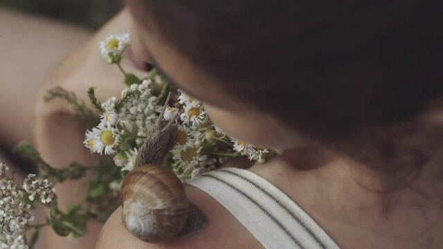 Concept of nature, cosmetology, spa, beauty. Woman holding bouquet of wild flowers or daisies in her hands. Close-up of snail slowly crawling along shoulder of young girl. Snail leaves muscus on skin.