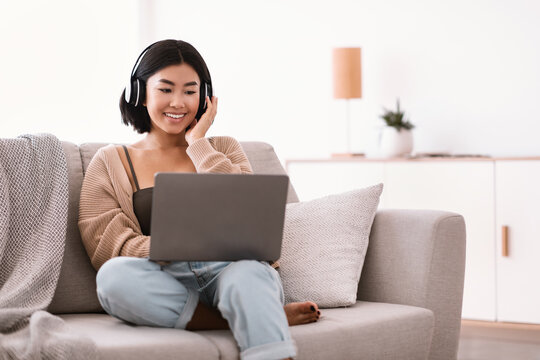 Asian lady listening to music wearing wireless headphones, using laptop
