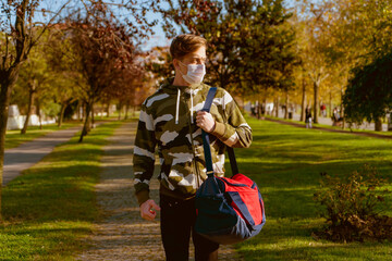 Obraz premium Young man going to the gym with his bag on his shoulder. wearing a protective mask is hiking through the woods. Young male athlete wearing a surgical mask is walking on the jogging track.