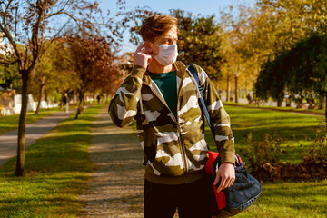Young man going to the gym with his bag on his shoulder. wearing a protective mask is hiking through the woods. Young male athlete wearing a surgical mask is walking on the jogging track.