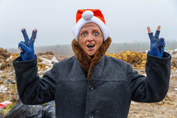 Obraz premium A dirty, poor woman in a New Year's hat, unsure of herself, shows two thumbs up against the background of a trash heap.