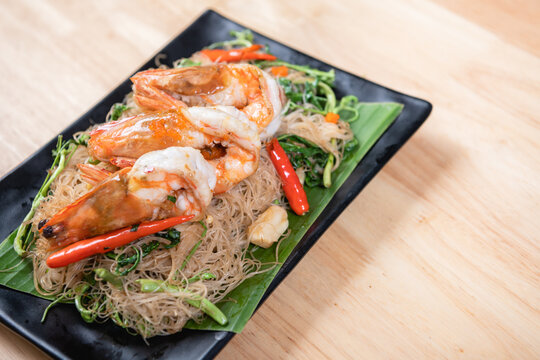 Shrimp, noodlesrice noodle and butter garlic fried shrimps sprinkled with pieces of chilli and chopped parsley on white dish on natural old wooden planks with spaghetti tongs, view from above, food
