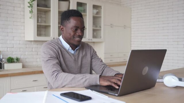 Smiling Professional Black African Business Man Using Laptop Working From Home Office. Happy African Male Customer Or Student Studying Online, E Learning, Typing On Computer Sits At Kitchen Table.