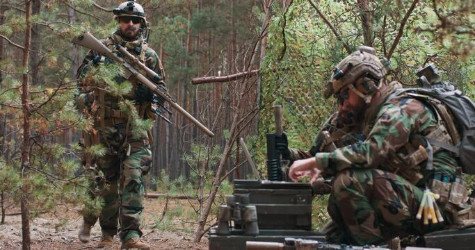Bearded soldiers in woodland military uniforms return from reconnaissance to a temporary base in the forest. In the foreground you can see the commander working on a laptop.