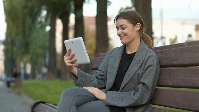 Young girl sits on the bench in autumn city square and talks via tablet computer