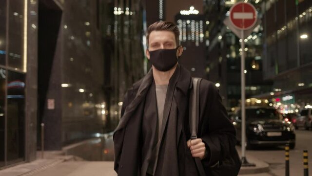 Businessman in black protective mask walk on background of business buildings, lights on. Gimbal night shot of caucasian man coming back from work