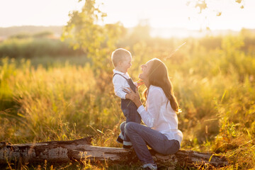 beautiful sunny photo of motherhood, Caucasian mother with long hair in summer at sunset plays with her son by log, in tall grass. Back light, soft focus and toning