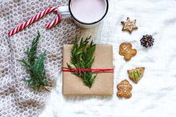 Eine Geschenk verpackt mit roter Schnur, eine Tasse heiße Schokolade und selbstgemachte Lebkuchen Kekse auf einer weißen Bettdecke. Hygge, Weihnachten, Vorfreude.