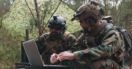 Bearded soldiers in uniform sit on military transport crates, analyze data on a laptop and work out tactics at a temporary forest base. In the background, you can see a soldier protecting the base. - Powered by Adobe