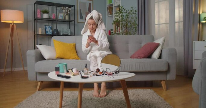 Middle plan of woman in bathrobe and towel sitting on sofa in living room texting, chatting, reading messages, smiling using smartphone and put her legs on couch. Makeup kits on table in living room.