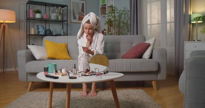 Middle plan of sad dissapointed woman in bathrobe and towel sitting on sofa in living room texting, chatting, reading and answering messages using smartphone. Makeup kits on table in living room.