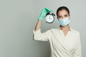 Serious doctor woman in medical protective face mask and professional uniform holding alarm clock...