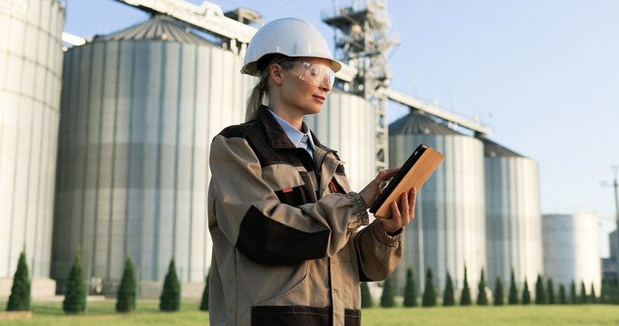 Beautiful joyful female engineer working on laptop and looking away. Portrait of pretty happy woman worker in helmet and goggles texting on computer outdoors at factory yard. Engineering concept