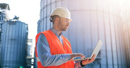 Side view of handsome Caucasian male worker in helmet and goggles typing on laptop while standing...