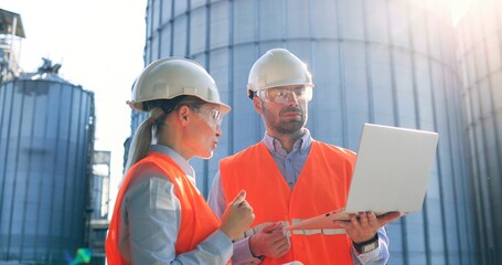 Male and female Caucasian engineers in helmets standing outdoors at plant near big tanks with...