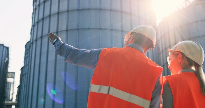 Rear of male and female Caucasian engineers in helmets and uniform standing outdoors at plant near big cisterns and talking. Woman and man controllers looking at away and discuss industry project