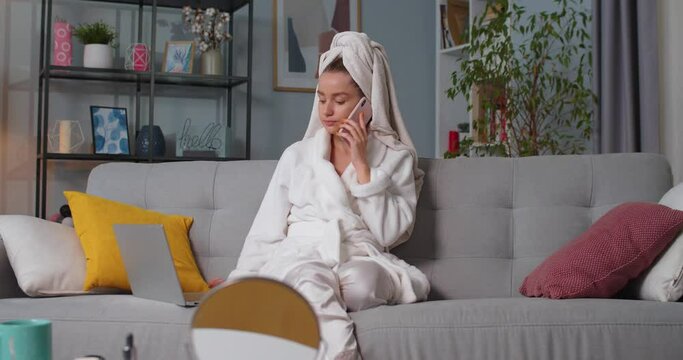 Close up of female in bathrobe sitting on sofa, finish speaking, talking on smartphone while typing on computer. Makeup kits on table in living room. Concept of lifestyle, self-care and wellness.