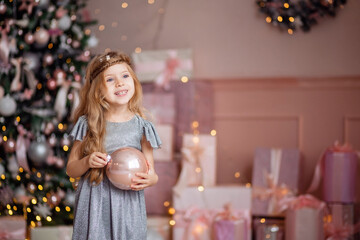 Cute little laughing blonde girl with long hair plays with a Christmas ball on a background of Golden yellow bokeh .