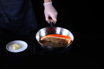 The cook fries a piece of red fish in a pan. Butter on a plate. Unrecognizable person. Photo on a black background. Top view.