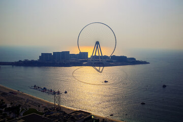 Dubai eye, UAE - 08.18.2020: Ferris wheel and Bluewaters island in backlight, sunset, with sun setting behind and reflection at sea surface