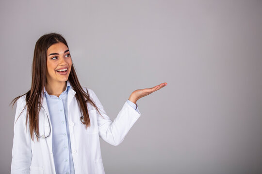 Cheerful young female doctor with stethoscope over neck looking at camera isolated on gray. Beautiful smiling woman doctor portrait in studio she is confident mood, she is pointing the right side