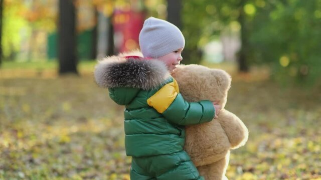 little boy playing with a plush toy in the park. funny baby otty in a warm down jacket and his teddy bear. close up view. Slow motion video. stock footage
