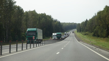 Fototapeta na wymiar Semi trucks lorry convoy move on suburban asphalted highway road on fence and green forest on roadsides background front view at summer day, on green forest background