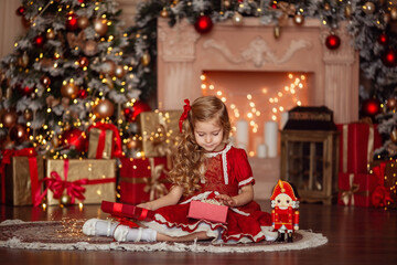 Happy cute little blonde girl with long hair in a red dress, waiting for Christmas and New year opens a gift on the background of bokeh and Christmas trees.
