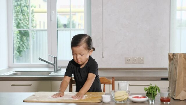 Pretty diligent toddler standing on chair to get cooking surface pours flour into wooden cutting board using hands and tablespoon