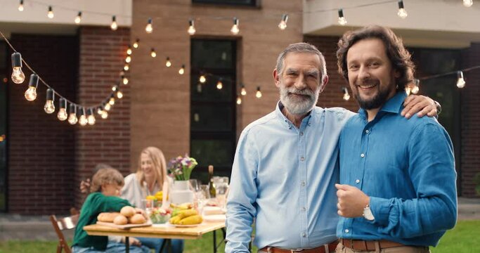 Portrait of handsome Caucasian two men, old and young, standing in hugs at back yard of house. Family dinner outdoor on background. Senior father with adult son embracing. Dad pensioner. Generations.