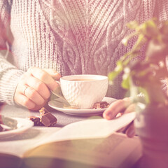 Coffee break composition with cup of coffee and chocolate. Woman drinking coffee and reading book. Toned photo with bokeh
