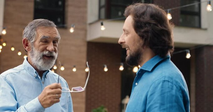 Handsome Caucasian two men, old and young, standing at back yard of house and talking. Family dinner outdoor on background. Senior father with adult son having talk and conversation. Generations.