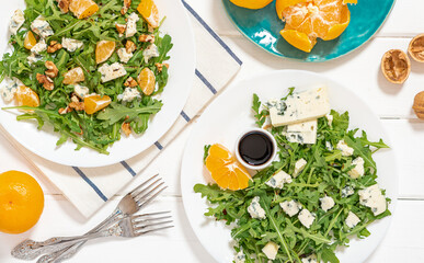 Arugula salad with gorgonzola cheese, tangerines, walnuts, and balsamic vinegar. Arugula and cheese salad in white plates on a white background top view. Light and healthy food.