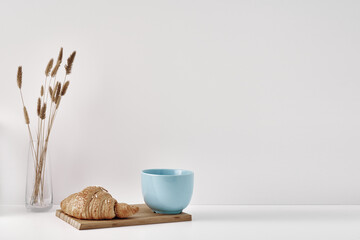 Croissant and a mug on a wooden stand, a vase and a pot with a plant on the books, white background. Eco-friendly materials in interior decor, minimalism. Copy space, mock up.