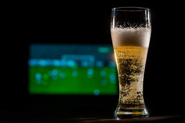 Close-up of fresh foaming light beer in a glass and football on TV on background. Sports bar concept