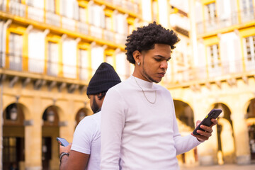 Lifestyle, two black Latino friends sending messages or watching social networks on their mobile. Boy with afro hair and boy with hat and earphones