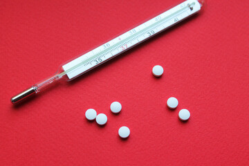 A mercury glass thermometer and seven small round tablets lie on a bright red surface. The thermometer lies with the numbers up. Treatment. Medicines.