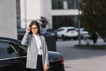 Fototapeta premium businesswoman touching eyeglasses while standing near black car