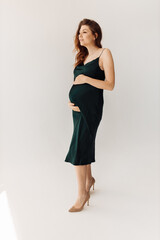 Wonderful pregnant woman stand in studio, posing, wearing green dress, expect for a first baby, happy lady excited about maternity, enjoy pregnancy, motherhood concept