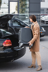  woman in stylish autumn clothes loading suitcase in car trunk