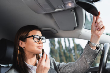 Fototapeta na wymiar businesswoman holding lipstick while adjusting rearview mirror in car