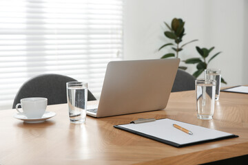 Modern laptop, glasses of water and clipboard on wooden table in conference room. Interior design