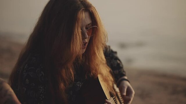 Red-haired hipster girl in beautiful sunglasses plays the guitar. Pretty female sings a song on sunset background. Vacation at the sea. Atmosphere of love. Self isolation, travel concept.